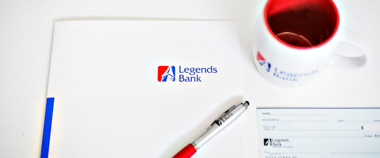 A photo of a desk with a Legends Bank folder, pen, check, and cup.